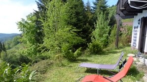 Un espacio al aire libre con césped, una mesa, dos sillas, una casa con ventana visible y abundante vegetación en el fondo.