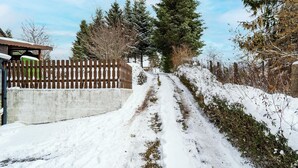 En snedækket sti der fører til en træport, omgivet af træer og en bygning med et skråt tag.