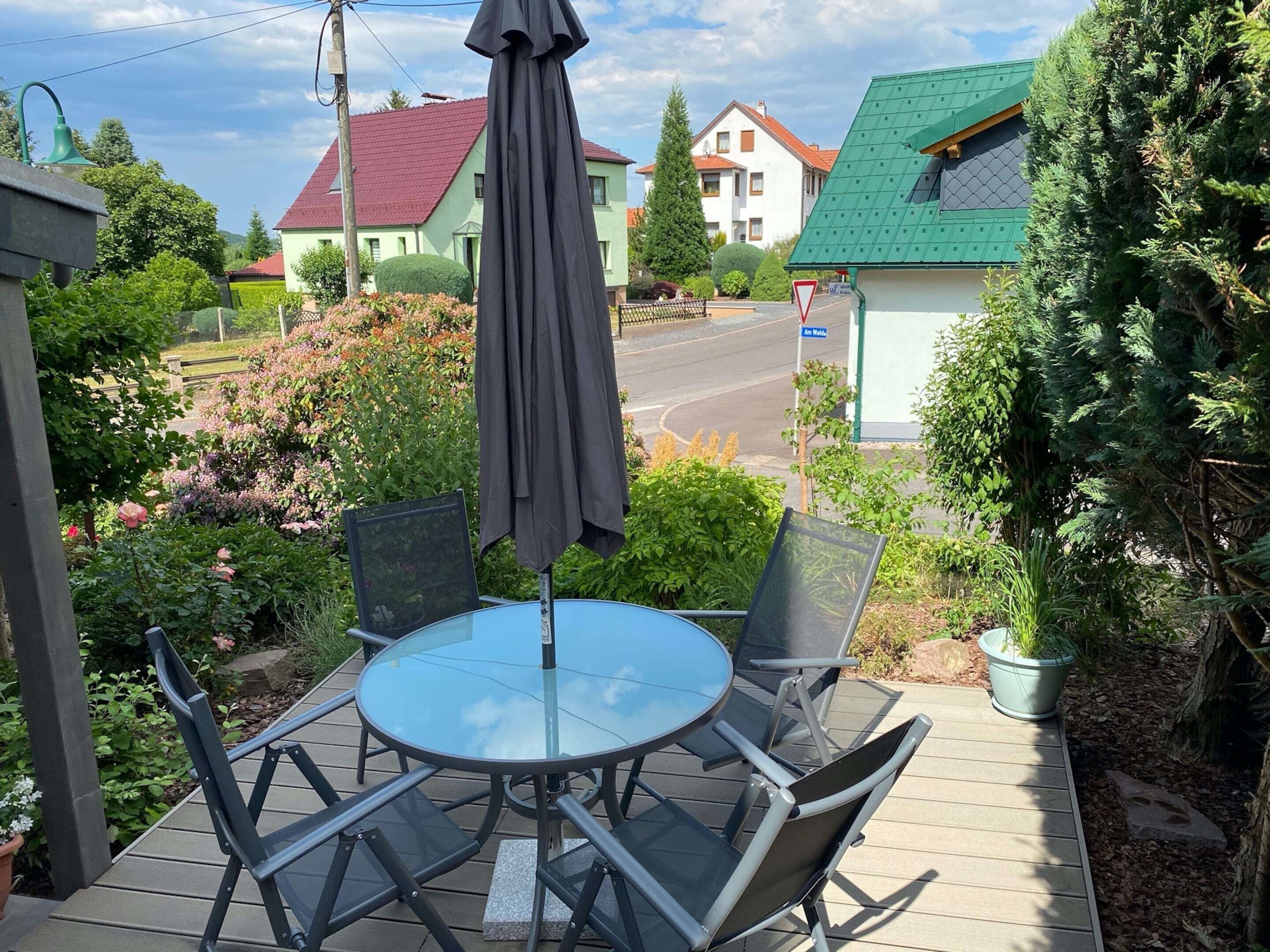 A patio with a round table and chairs, surrounded by greenery and a street view.