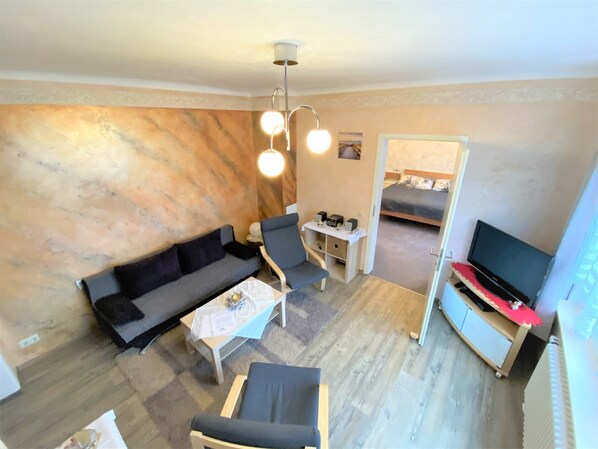 Un espacio habitable pequeño con un sofá, una mesita, una silla, un televisor y una cama al fondo.