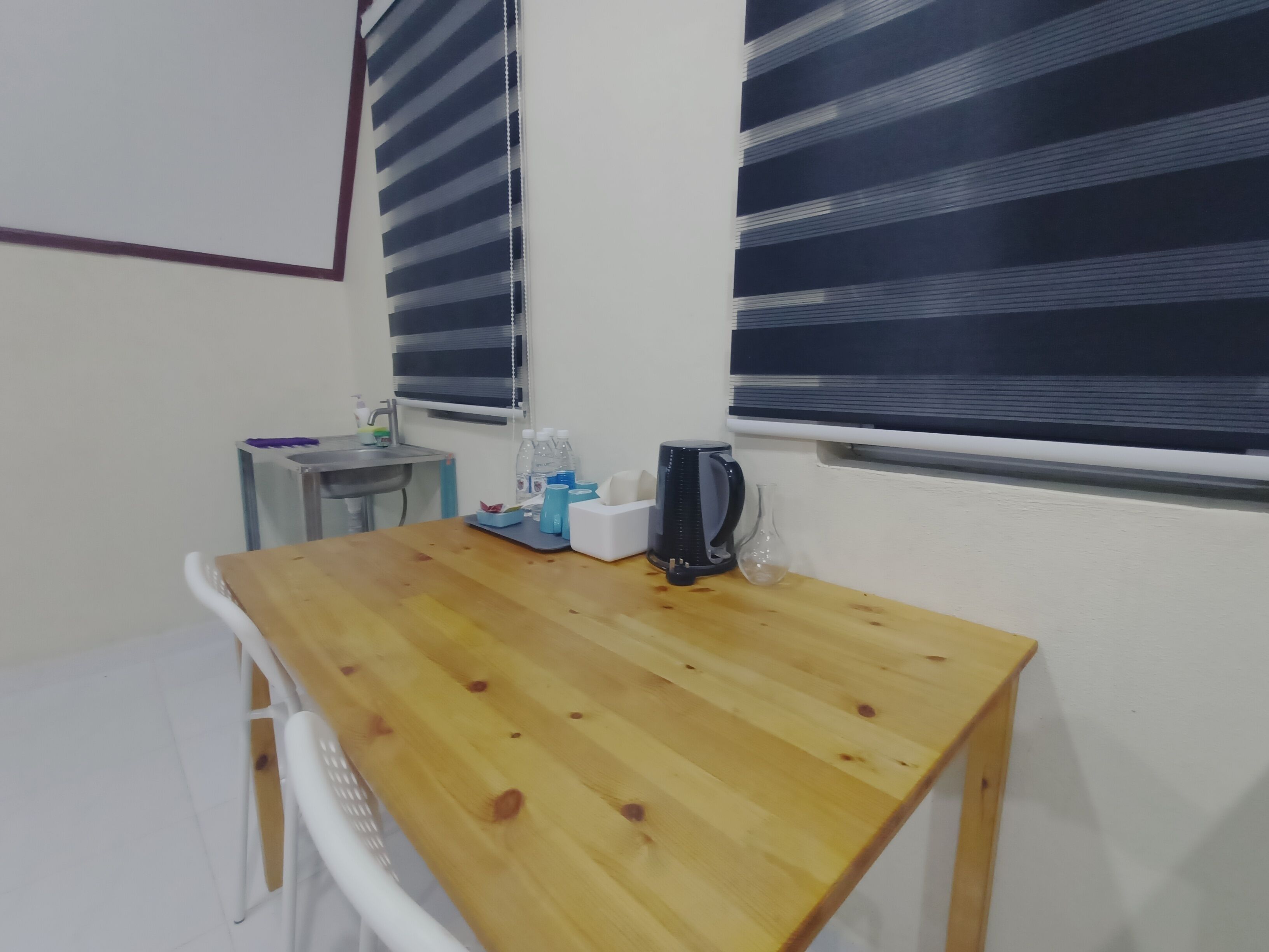 Ein kleiner Holztisch mit einem Wasserkocher, einer Taschentuchbox und einer Wasserflasche. Ein weißer Stuhl und ein kleiner Tisch mit einem Waschbecken und einer Handseifenflasche stehen daneben.