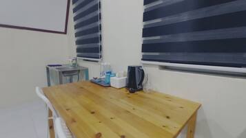 Ein kleiner Holztisch mit einem Wasserkocher, einer Taschentuchbox und einer Wasserflasche. Ein weißer Stuhl und ein kleiner Tisch mit einem Waschbecken und einer Handseifenflasche stehen daneben.