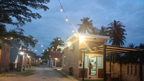 Un camino lleva a un edificio con un letrero que indica que está abierto. Hay luces colgantes, palmeras al fondo y una zona techada con mesas.
