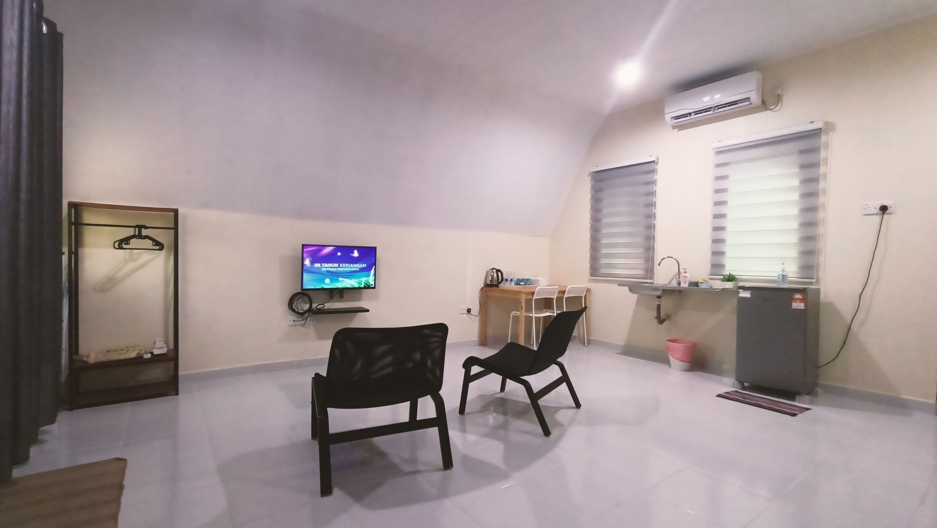 Een kleine, goed verlichte kamer met een flatscreen televisie, twee zwarte stoelen, een houten bureau, een klein wastafeltje en een airconditioning.'