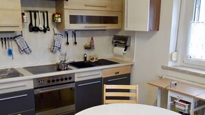 Een kleine keuken met een fornuis, oven en gootsteen. Er staat een houten stoel bij een tafel met een witte tafelkleed.