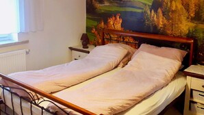 Een bed met een houten hoofdbord, een nachtkastje met een lamp, en een muur met een schilderij van een berglandschap.