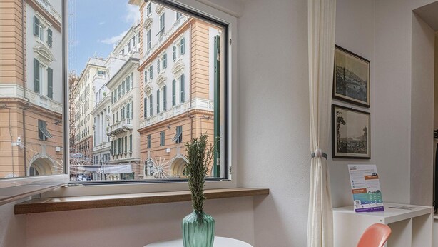 Een hotelkamer met een groot raam dat uitzicht geeft op historische gebouwen, een tafeltje met een plant en een bureau met een computer en een stoel.