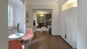 Een smalle gang met een eethoek, bestaande uit een kleine tafel en twee stoelen, een keuken met kasten en een aanrecht, en een deur naar een andere kamer.