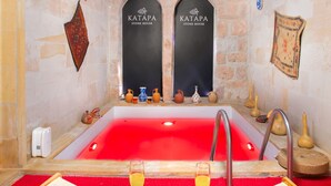 A stone house interior with a red bathtub, two black doors labeled 'Katapa Stone House', and a tray with two glasses of yellow liquid. - Katapa Stone House (Nevsehir)
