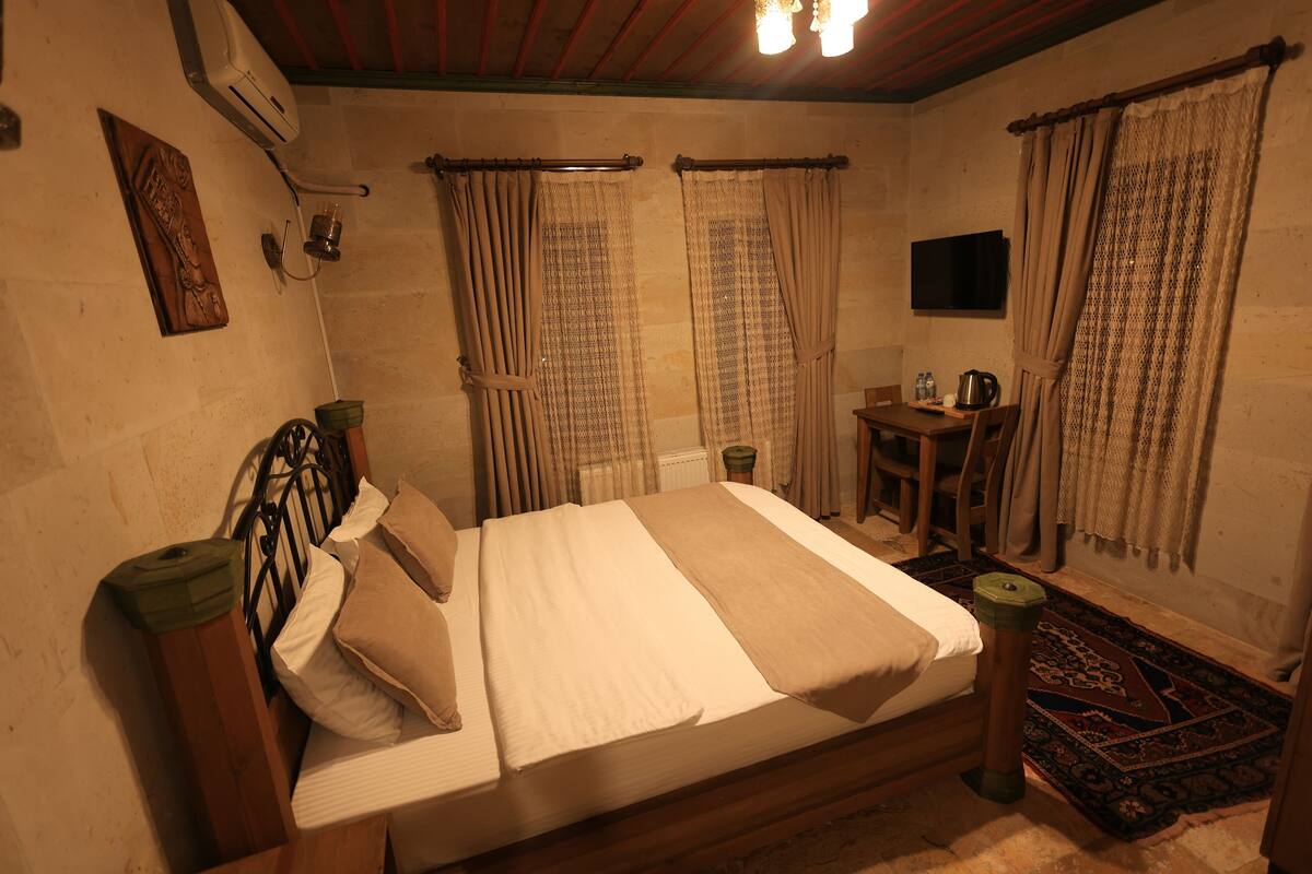 A bedroom with a bed, a small table with a kettle, curtains, and a wall-mounted TV.