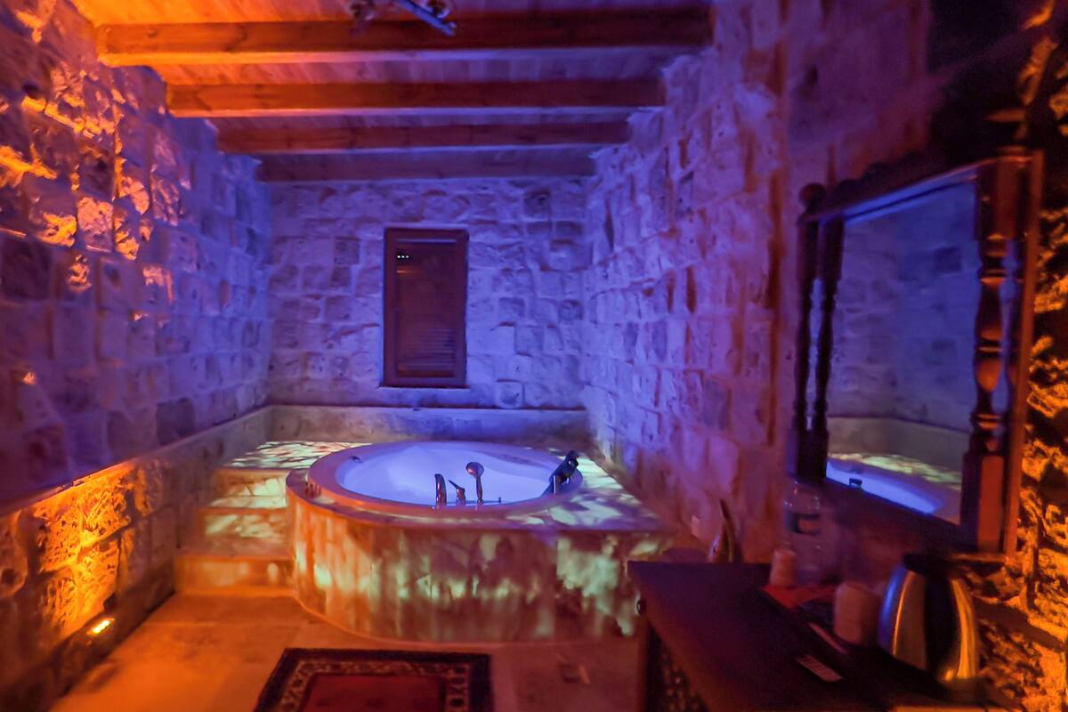 A stone-walled bathroom with a central bathtub, a wooden bench, and a mirror.