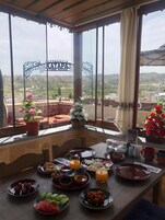 Mesa de comedor dispuesta con diferentes platos y bebidas, rodeada de amplias ventanas con vistas a un paisaje montañoso.