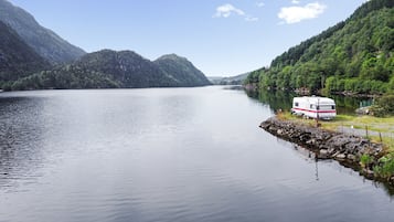 Una caravana blanca estacionada frente a un lago tranquilo, con montañas al fondo.