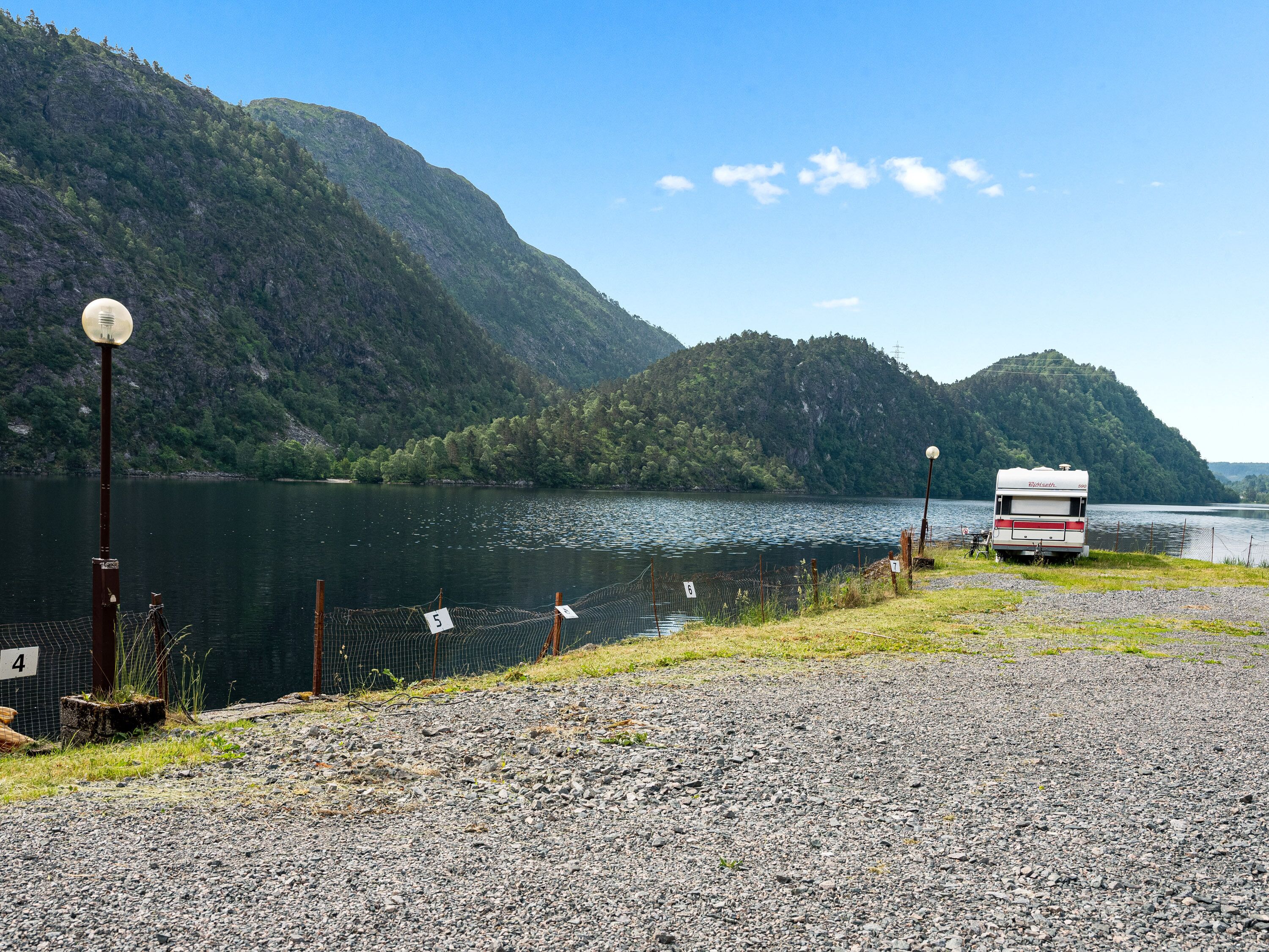 Ein Kiesbereich mit einem See, einem bewaldeten Berg und einem weißen Wohnmobil am Wasser.