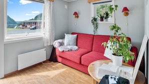 Un salon moderne avec un canapé rouge, un plancher en bois, une fenêtre donnant sur une maison et des montagnes, ainsi que plusieurs plantes en pot.