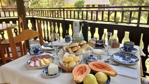 A table set with various food items and drinks, including bread, pastries, and a fruit bowl, on an outdoor deck with a railing. - Pousada Lago Azul  (Camanducaia)