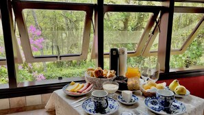 A breakfast table with various food items and a thermos, set against a backdrop of a forest view through large windows. - Pousada Lago Azul  (Camanducaia)