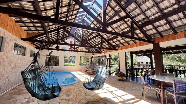 A covered outdoor area with a pool, hanging chairs, and a table with chairs. - Pousada Lago Azul  (Camanducaia)