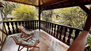 A covered balcony with two wooden chairs and a view of a garden and a small building. - Pousada Lago Azul  (Camanducaia)