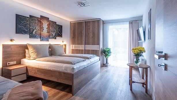 Quarto moderno com cama de madeira, mesas de cabeceira, uma pequena mesa de cabeceira com um vaso de flores e uma janela grande com vista para o exterior.
