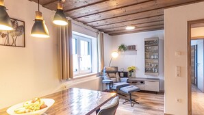 Uma sala de jantar moderna com uma mesa de madeira, cadeiras e uma cesta de frutas. Há uma janela com cortinas, um radiador e uma estante com copos.