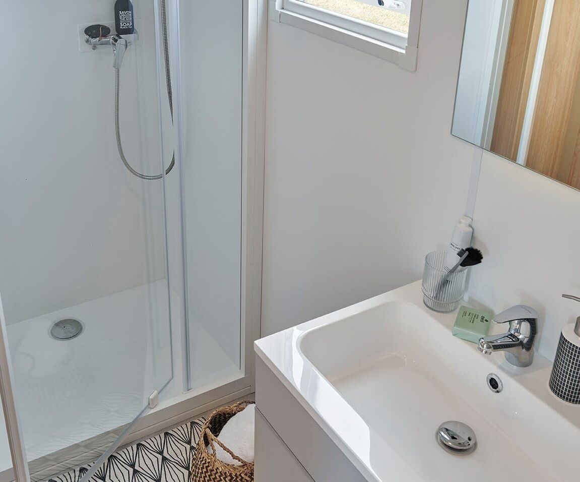 Un baño con ducha de vidrio, un lavamanos blanco con espejo y una ventana pequeña.