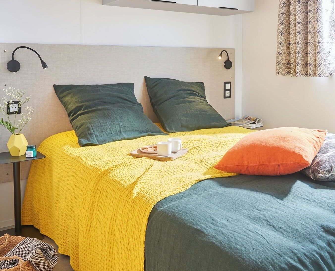 Una cama bien hecha con un edredón amarillo, dos almohadas verdes y una almohada naranja. Hay una mesita con un florero y una bandeja con una taza y su platillo.