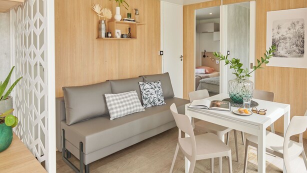 A modern living room with a grey sofa, white chairs, a wooden wall, and a dining table with a vase and books.
