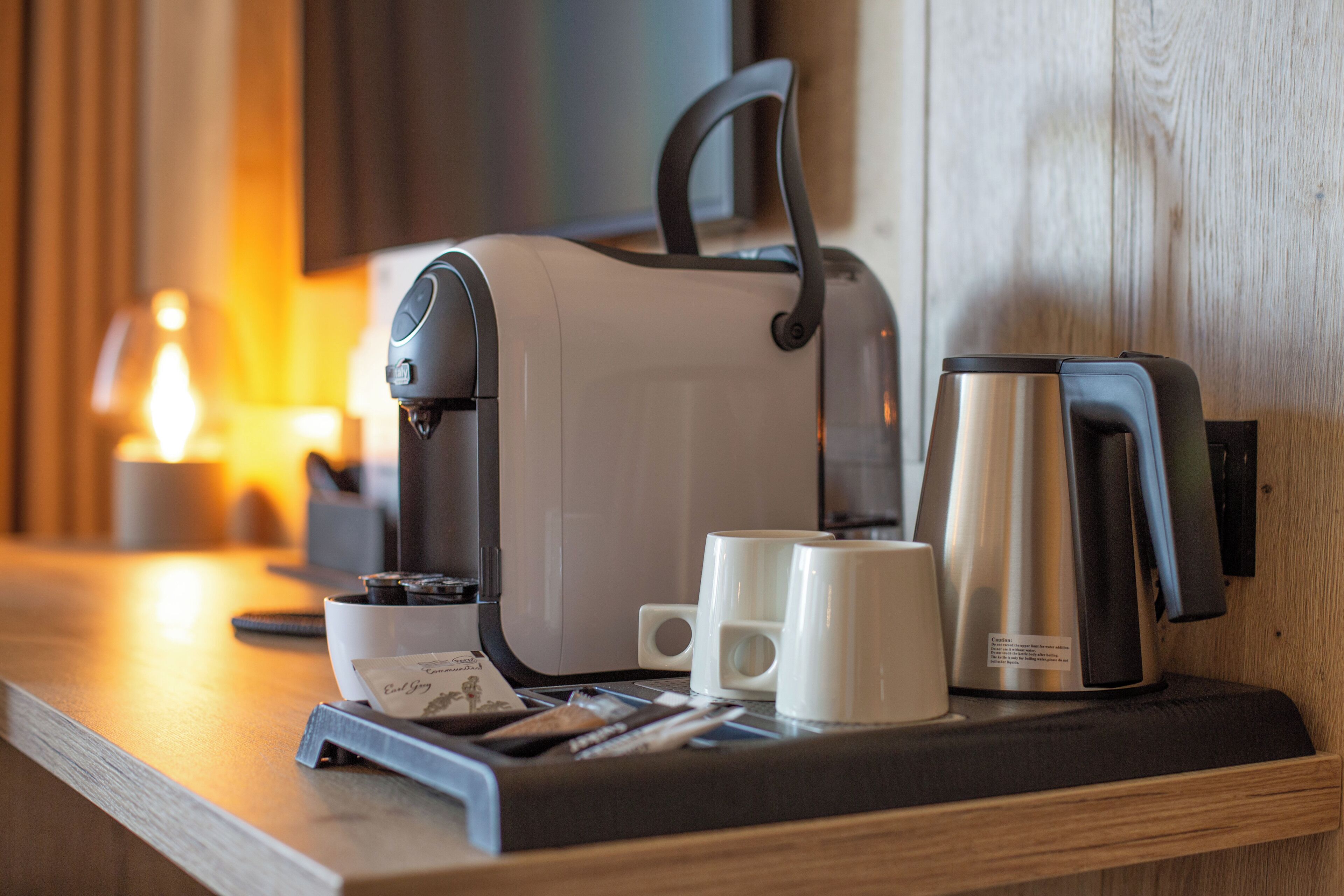 A coffee maker, two white cups, a tray, and a small candle on a wooden counter.