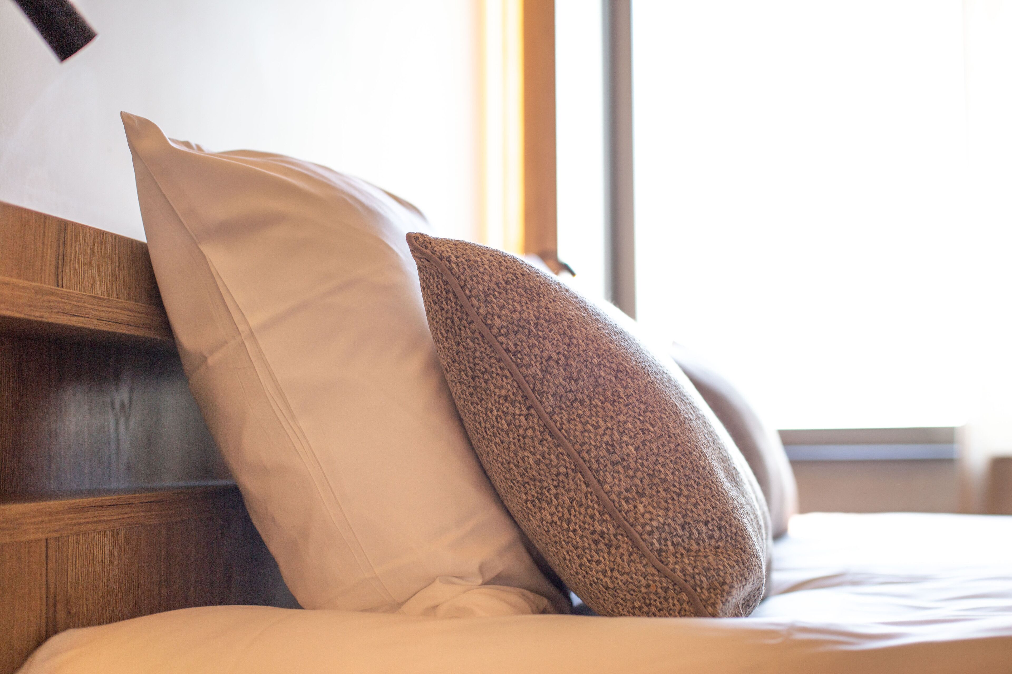 A bed with two pillows, one plain and one textured, in a room with a wooden headboard and a window.