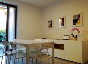 Un comedor con una mesa y sillas blancas, una pequeña barra de cocina y una orquídea en maceta.