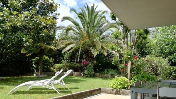 Un espace patio avec une chaise en métal blanc, une table et une table avec un plateau en verre. On aperçoit des palmiers et diverses plantes en arrière-plan.