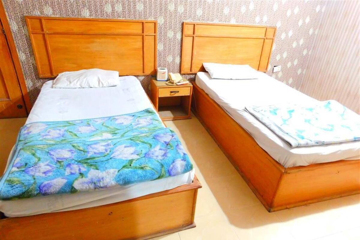 A room with two wooden beds, each with a nightstand. The beds have white linens and one has a floral bedspread. The room has patterned wallpaper and a tiled floor.