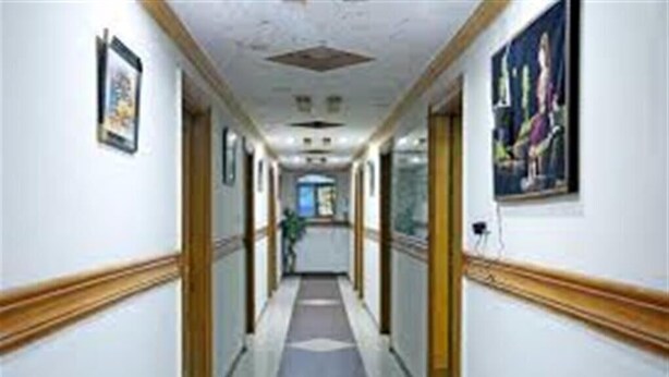 A long, narrow hallway with white walls and wooden trim. There are framed pictures on the walls and a painting on the right side. The floor is tiled. - Hotel Al Noor (Lahore)