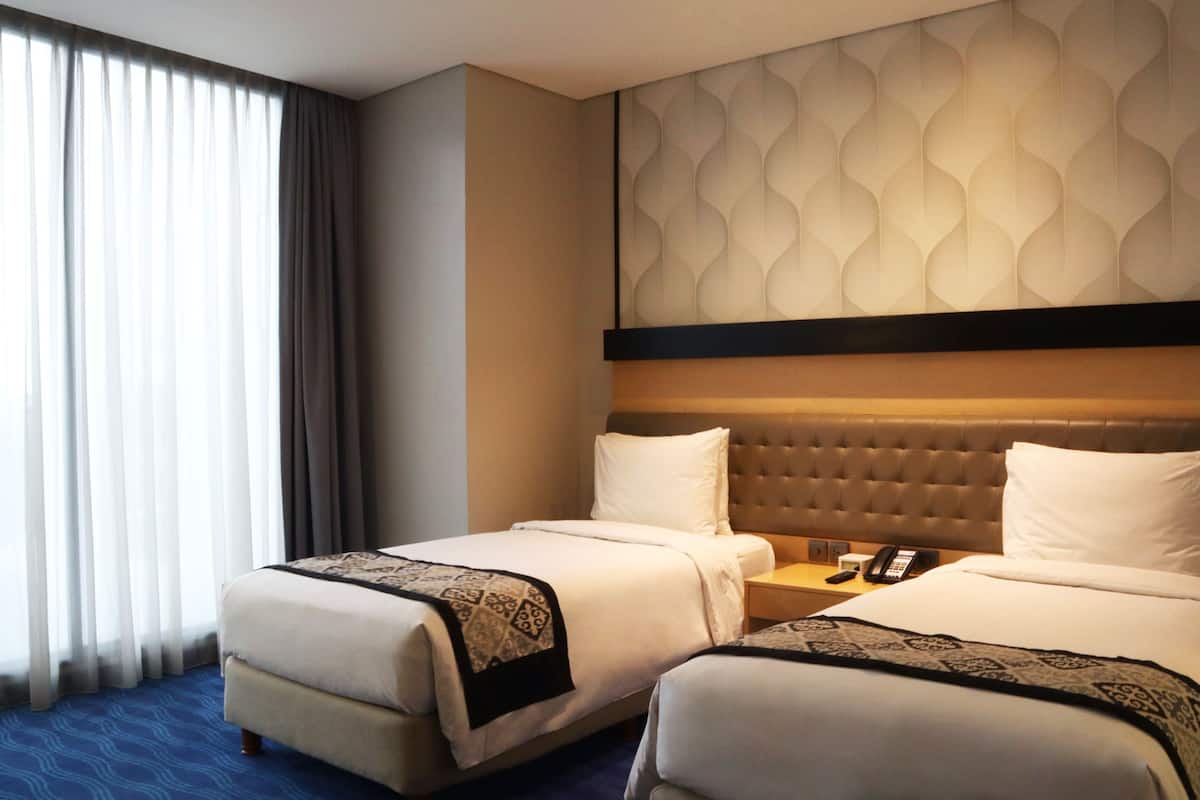 A hotel room with two beds, a large window with curtains, a patterned wall, and a telephone on the nightstand.