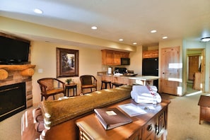 A living room with a fireplace, a television, and a sofa. There is a kitchen area with wooden cabinets and a microwave.
