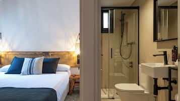Habitación de hotel con cama, mesita de noche y zona de ducha con paredes de vidrio.