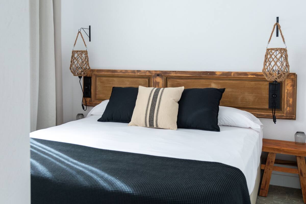 A neatly made bed with a wooden headboard, a bedside table with a lamp, and a hanging light fixture.