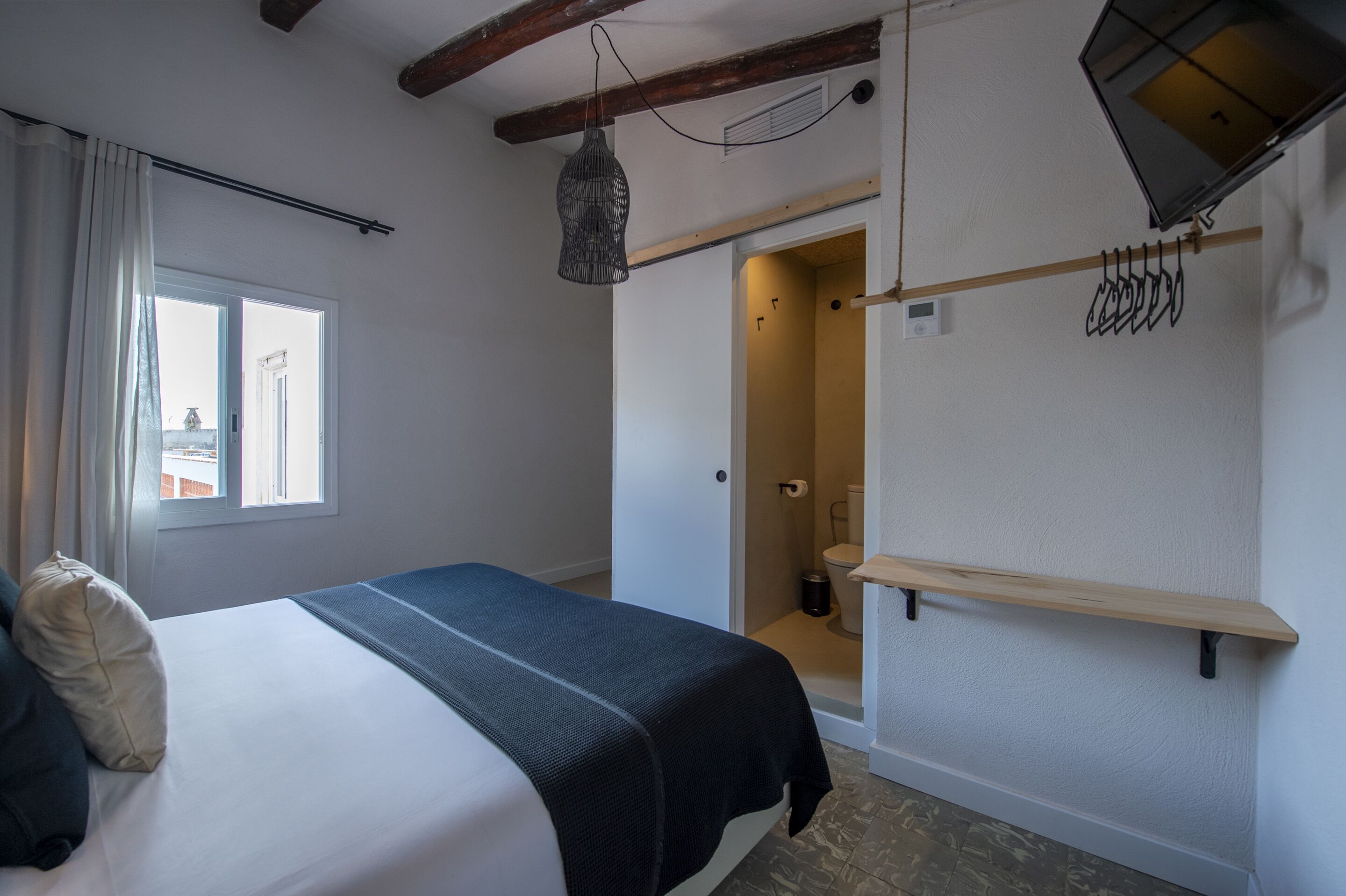 A bedroom with a bed, a window, a hanging light fixture, a wooden shelf, and a door leading to a bathroom.