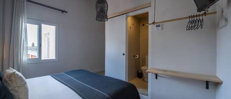 A bedroom with a bed, a window, a hanging light fixture, a wooden shelf, and a door leading to a bathroom.