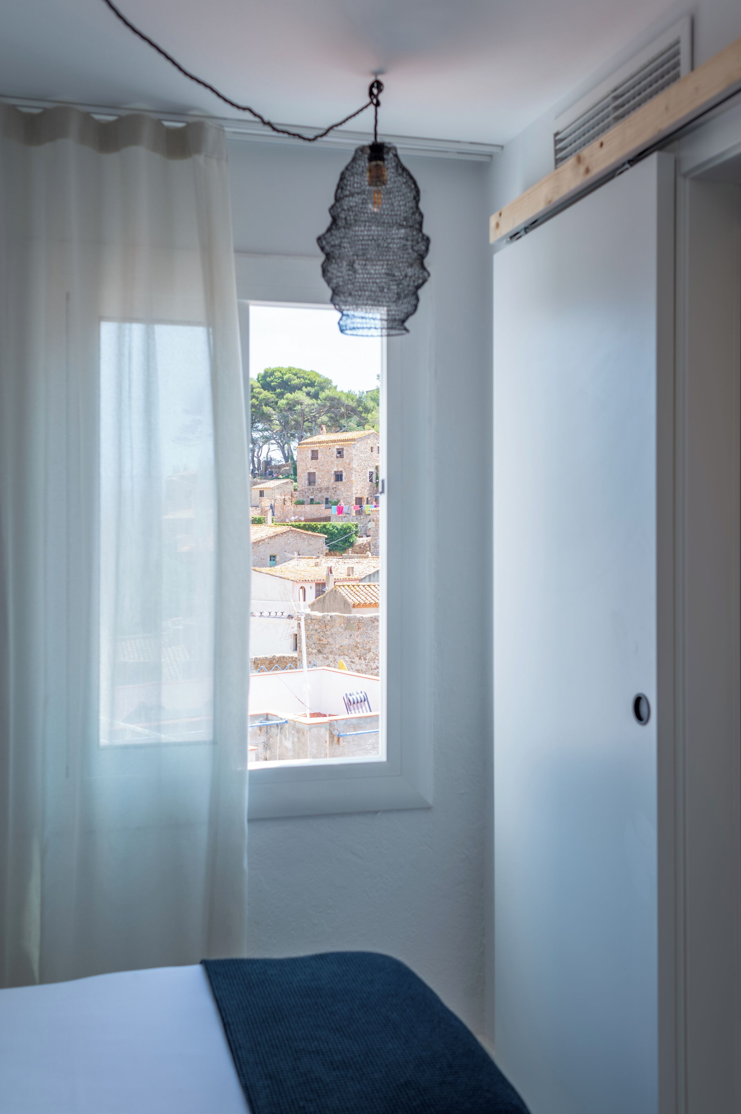 Ein Schlafzimmer mit einem Bett, einem Fenster mit Blick auf Gebäude und einer hängenden Leuchte.
