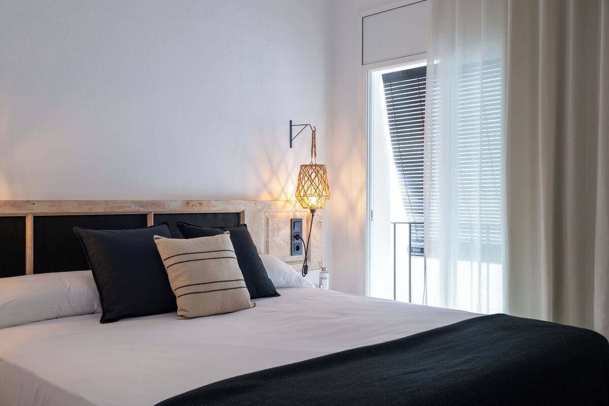 A neatly made bed with a wooden headboard, a bedside lamp, and a window with blinds.