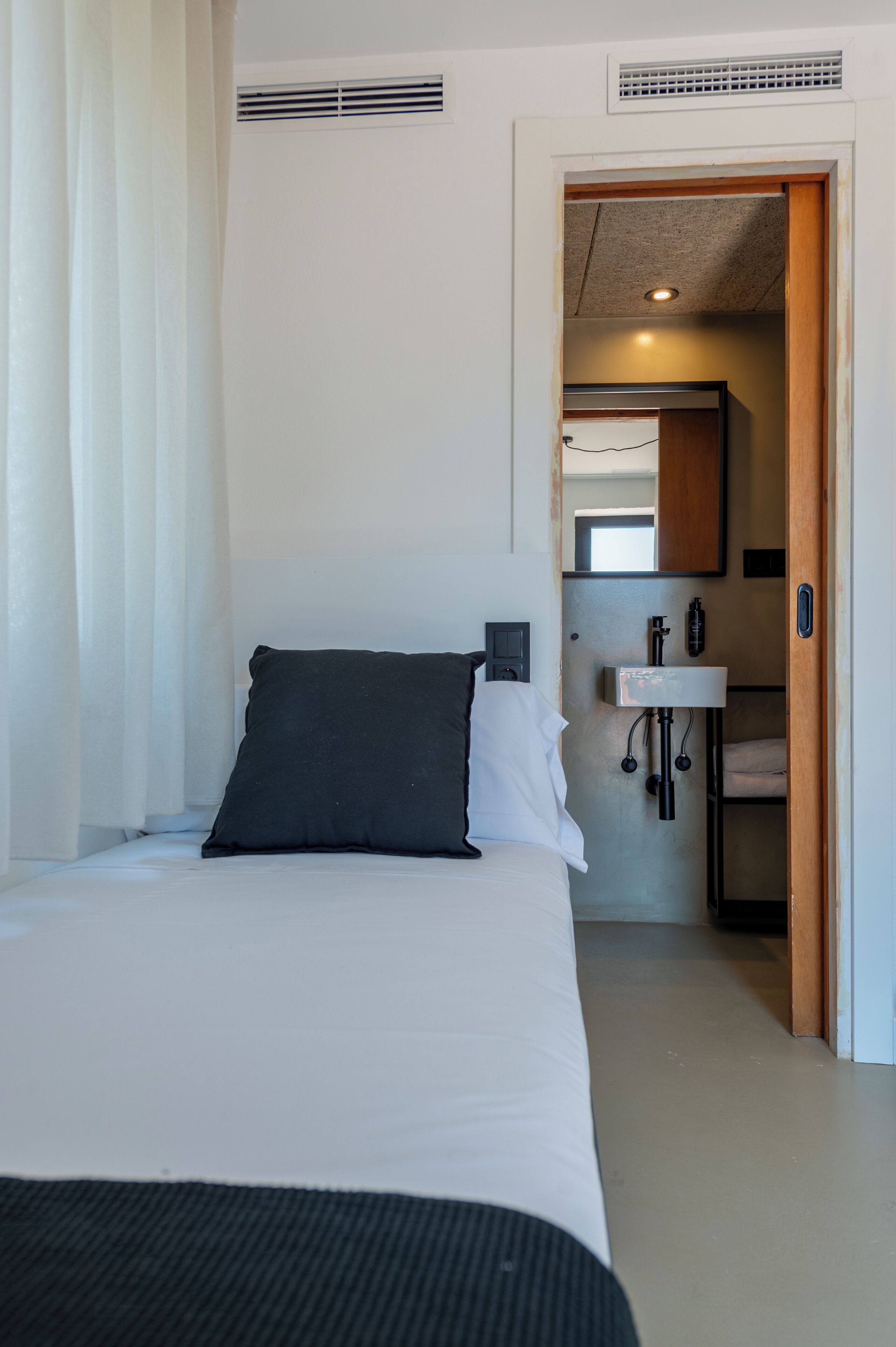 A bedroom with a bed, a bedside table, a wall-mounted light, a window with curtains, and a bathroom with a sink and mirror.