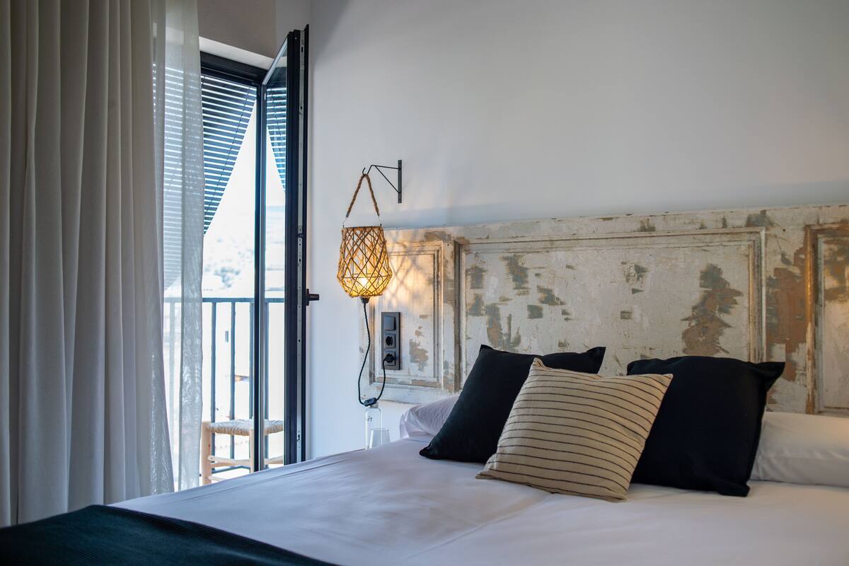 A hotel room with a bed, a window with blinds, a hanging light fixture, and a view of the outside.