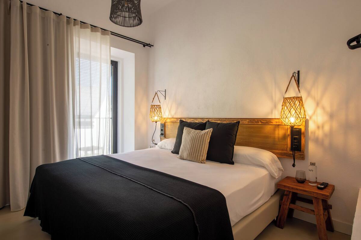 A neatly made bed with a black bedspread and white linens, two wall-mounted lamps, a small wooden nightstand with a glass of water and a remote control, and a window with white curtains.