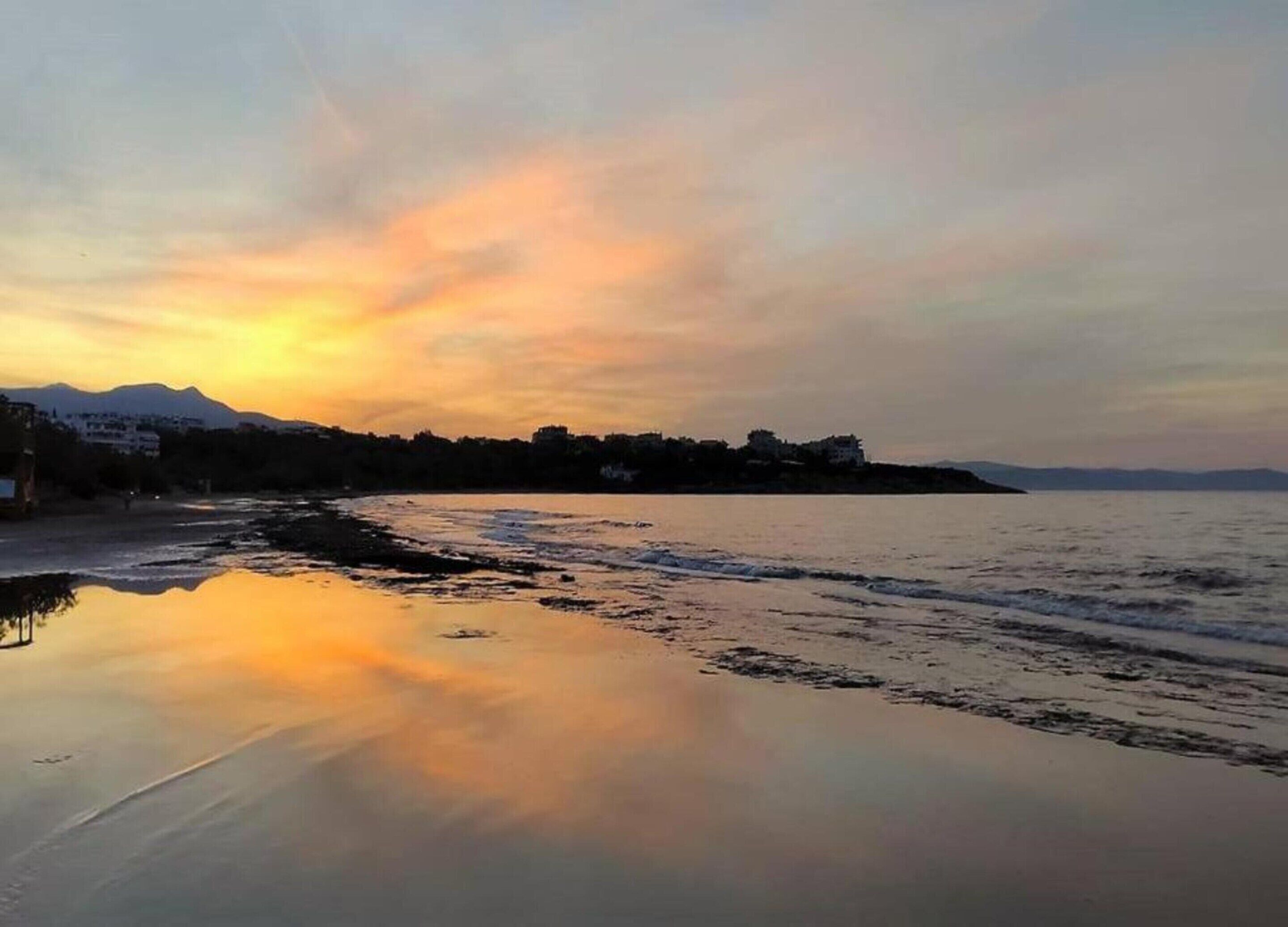 A coastal scene with a calm sea reflecting the sky, a sandy beach, and a distant mountain range.