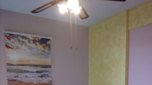 A neatly made bed with white linens and towels, a ceiling fan, a painting of a beach scene, and a yellow accent wall.