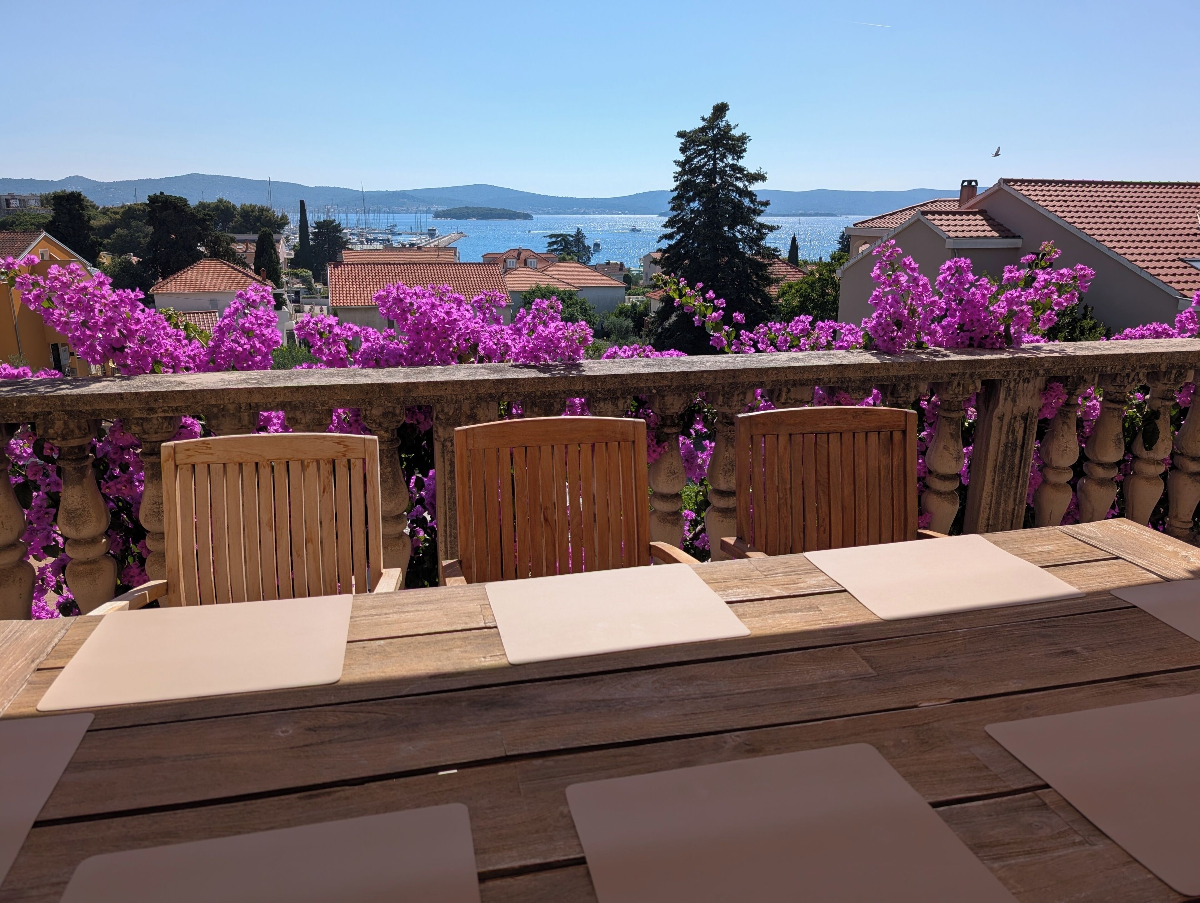 A terrace with wooden chairs and tables, overlooking a body of water and a distant mountain range.