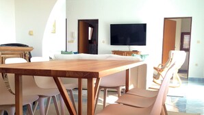 A modern living room with a wooden dining table, white chairs, a flat-screen TV, and an arched doorway.