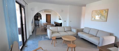 A spacious living area with a sofa set, a coffee table, and a dining table. An archway leads to a kitchen and dining area.